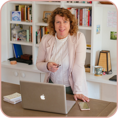 Photo of middle-aged white business woman with shoulder-length medium hair leaning on her left hand, which is resting on a home desk with a computer, phone and notebook. She is wearing a light pink jersey jacket, a feminine white blouse and holding a pair of glasses in her right hand. Behind her are white bookshelves in a built-in unit and there is a cup of tea next to a lamp on the right hand side of the unit.