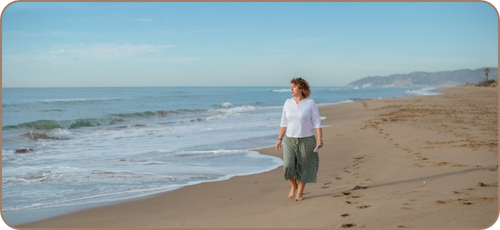 Middle-aged white woman with shoulder-length curly hair in a green skirt and white three-quarter-sleeve t-shirt and holding a small notebook walking barefoot along a sandy beach looking to her right at the white surf and blue sea. There are low mountains and a lone palm tree in the far distance in the upper right quadrant of the image. There are no other people on the beach.