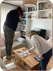 White female photographer standing on a low stool, leaning over to take a picture of 6 books arranged on a table and another white woman is leaning to touch one of the books.
