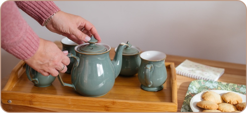 Photo of a white woman's hands on a green teapot with matching mugs and sugar bowl on a wooden tray. There is a plate of biscuits on a green tray and a notebook on the table as well.