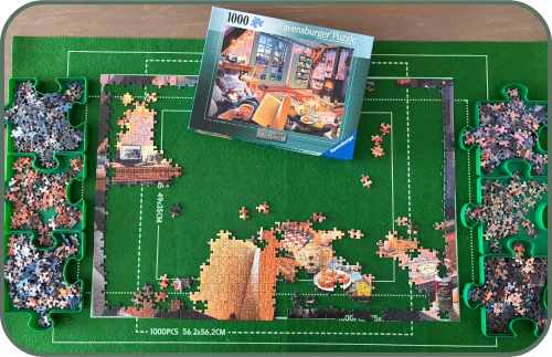 Picture of a partially completed jigsaw puzzle with the rest of the pieces separated neatly into 6 puzzle-shaped trays. The puzzle box shows the full picture of a cosy living room and in the foreground a woman's hands are holding an open book and a mug of tea.