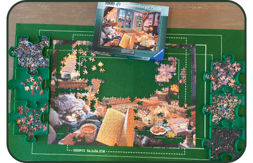 Picture of a half completed jigsaw puzzle with the rest of the pieces separated neatly into 6 puzzle-shaped trays. The puzzle box shows the full picture of a cosy living room and in the foreground a woman's hands are holding an open book and a mug of tea.