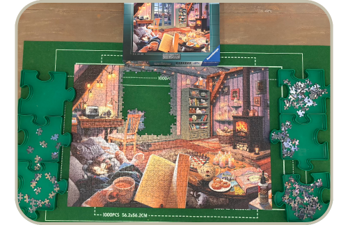 Picture of an almost complete jigsaw puzzle with the few remaining pieces separated neatly into 6 puzzle-shaped trays. The puzzle box shows the full picture of a cosy living room and in the foreground a woman's hands are holding an open book and a mug of tea.