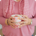 A woman's hands with a French manicure, silver rings, a silver watch and two light purple beaded bracelets holding a pink flowery mug with golden tea in it. Only the woman's torso is visible and she is wearing a medium-tone pink sweater and a matching scarf with tiny white stars on it.