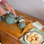 A woman's hands are pouring tea from a green Denby teapot into a matching green Denby mug on a tray with a matching sugar bowl. The tray is resting on a warm brown wood table with plate of homemade butter biscuits on a William Morris tray and a flowery notebook to the side of it.