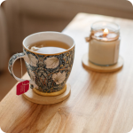 Herbal tea in a William Morris Pimpernel pattern mug with a lit candle in soft focus on a section of a light brown wood table