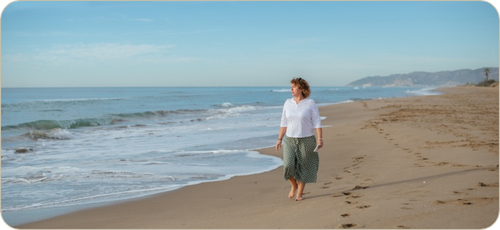 Middle-aged white woman with shoulder-length curly hair in a green skirt and white three-quarter-sleeve t-shirt and holding a small notebook walking barefoot along a sandy beach looking to her right at the white surf and blue sea. There are low mountains and a lone palm tree in the far distance in the upper right quadrant of the image. There are no other people on the beach.