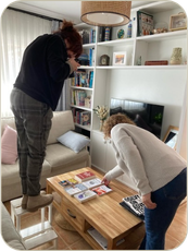 White female photographer standing on a low stool, leaning over to take a picture of 6 books arranged on a table and another white woman is leaning to touch one of the books.