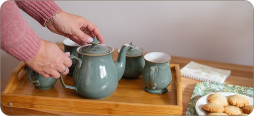 Photo of a white woman's hands on a green teapot with matching mugs and sugar bowl on a wooden tray. There is a plate of biscuits on a green tray and a notebook on the table as well.