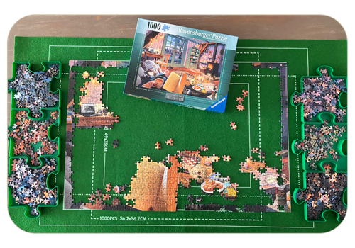 Picture of a partially completed jigsaw puzzle with the rest of the pieces separated neatly into 6 puzzle-shaped trays. The puzzle box shows the full picture of a cosy living room and in the foreground a woman's hands are holding an open book and a mug of tea.