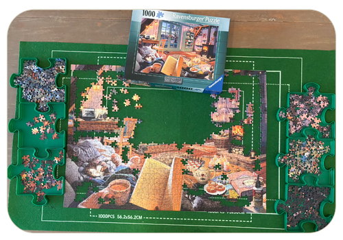 Picture of a half completed jigsaw puzzle with the rest of the pieces separated neatly into 6 puzzle-shaped trays. The puzzle box shows the full picture of a cosy living room and in the foreground a woman's hands are holding an open book and a mug of tea.